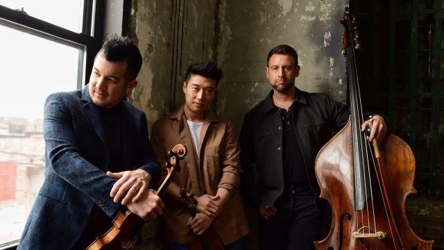 The three members of Time for Three pose for a picture next to a large picture window. They are wearing suits and holding their instruments. They are bathed in a soft light.